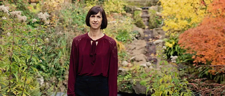 Landscape image of a woman stood in a green garden park looking direct to camera