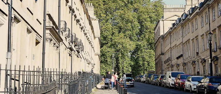 Landscape view along a residential property street with terraced houses either side and cars parked along the right. People walking down the street on the left