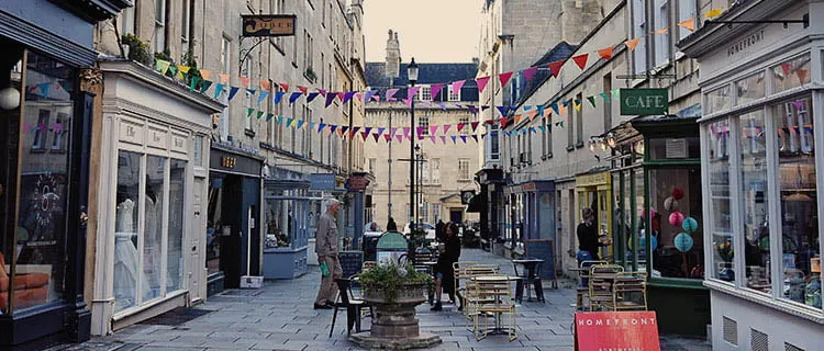 Landscape image of a pedestrianised street with commercial buildings bunting, seats and tables and cafes on either side.