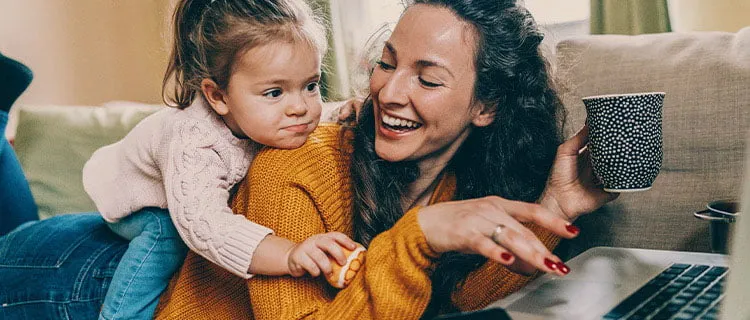 Landscape of woman on sofa holding mug of coffee in her hand with child climbing on her back.