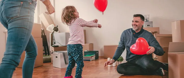 Man sat on floor in front of partner and child holding a heart shaped balloon with unpacked boxes behind him.