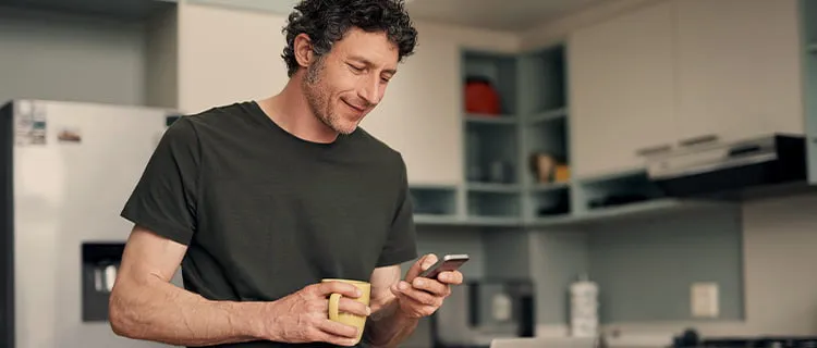 Landscape image of man in kitchen holding a mug of coffee and using iPhone