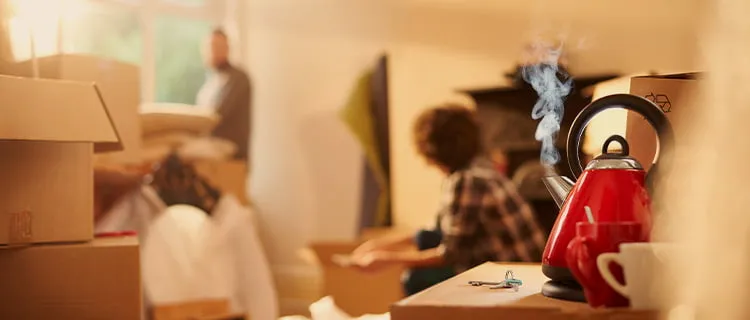 Landscape image of foreground focus of kettle boiled with steam from the spout, on top of cardboard boxes next to set of new keys. In the background is more unpacked boxes with two people talking.