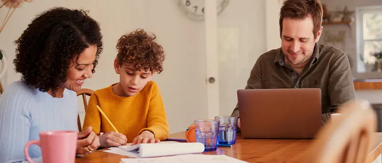 Image of family at kitchen table, father on laptop and mother and child are working together on a pad of paper.