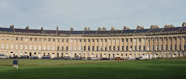 Wide angle shot of the Royal Crescent in bath with two people walking across the green field in the foreground.