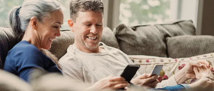 Woman and man sat on sofa, woman with feet up on the sofa. both looking at their phones and laughing.