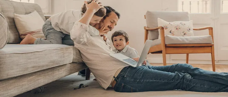 Landscape of man sat in front of sofa on the floor with laptop and two children besides him watching screen laughing.