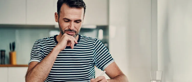 Man sat at kitchen worktop with pen in hand and hand on chin writing notes.