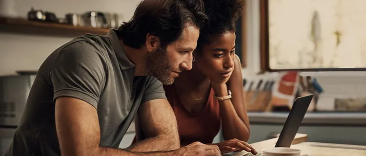 Landscape of two people sat hunched over a laptop screen on a desk in the kitchen, engaging with what's on the screen.