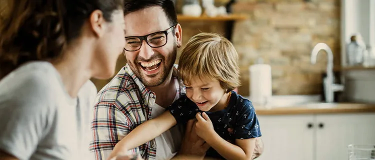 Landscape image of man holding child while joking and laughing with woman in foreground.