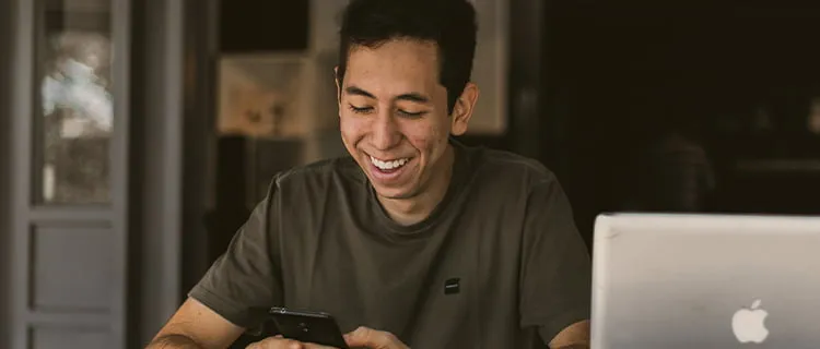 Landscape of man laughing at home on his mobile phone with laptop seen in bottom right hand corner.