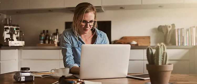 Landscape image of woman sat at kitchen room table with laptop and iPad working. In the foreground is pends, paper, camera and a cactus plant.