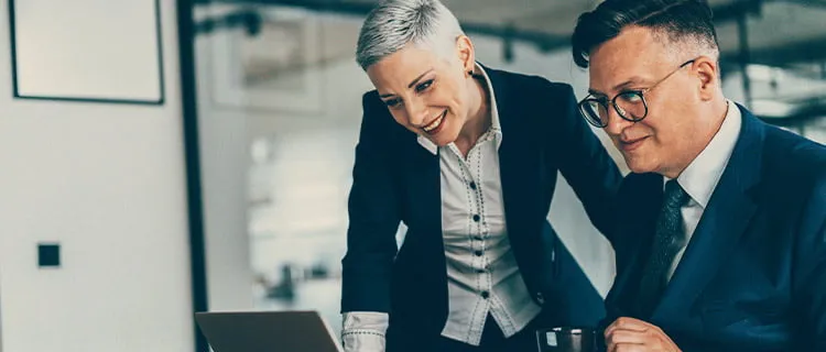 Image of two people in an office room, one is sat down the other stood up, both looking at a laptop screen.