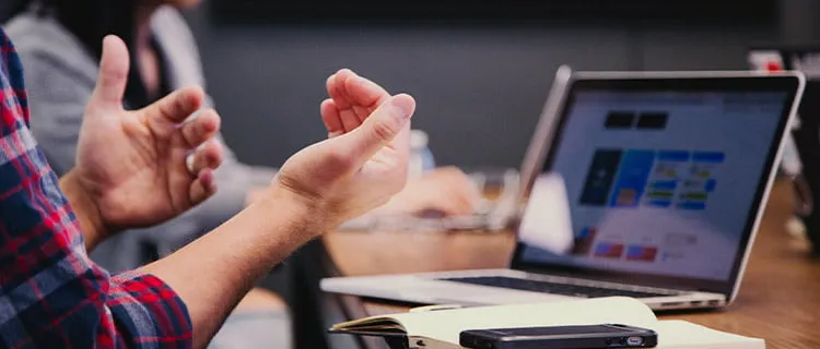 Image of a laptop screen with notepads and mobile phones in the foreground. People's heads out of shot but hand gestures can be seen indicating people are having discussion in a meeting room.