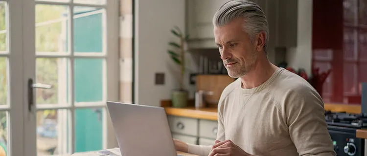 Landscape image of man sat at kitchen worktop working on laptop.