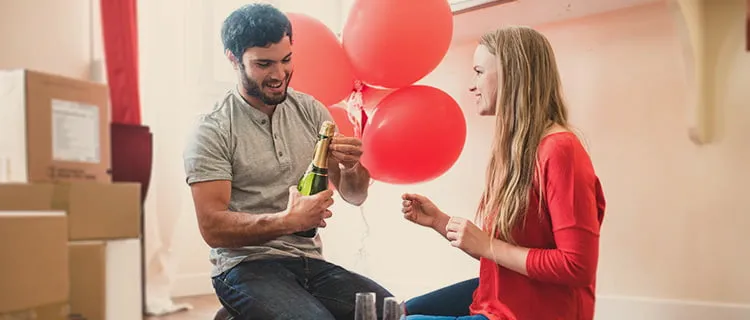 Landscape image of couple opening bottle of champagne, with balloons and boxes in the background