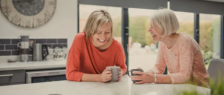 two women share a laugh while sat at home kitchen island drinking coffee