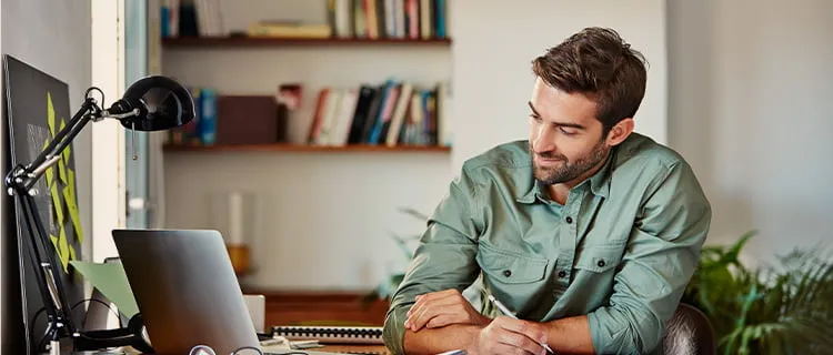 Man in green shirt in home office writing notes while looking at laptop screen, book shelves in the background.